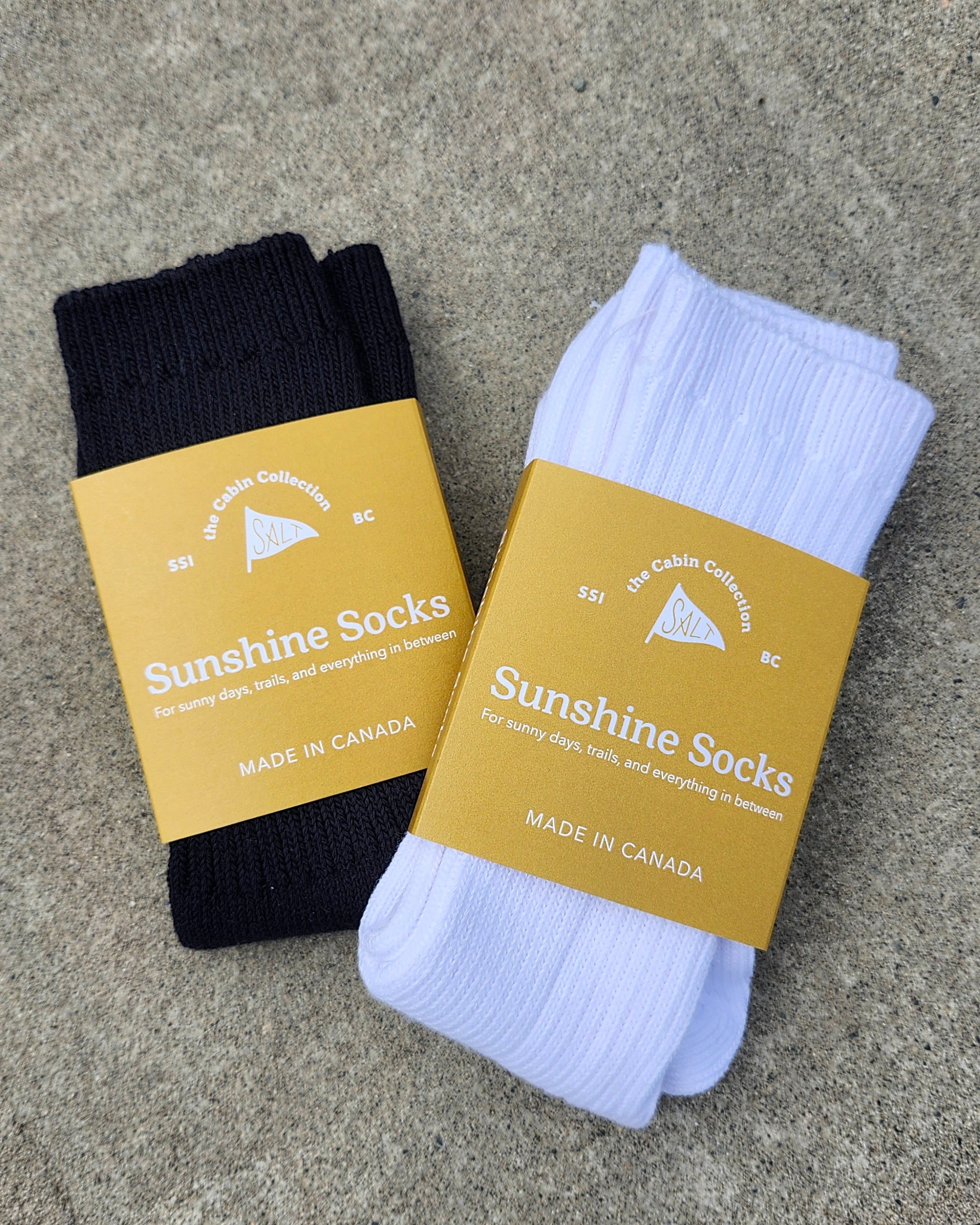 Two pairs of Sunshine Socks, one white and one navy, each wrapped in a mustard yellow paper band, displayed on a textured gray concrete surface.